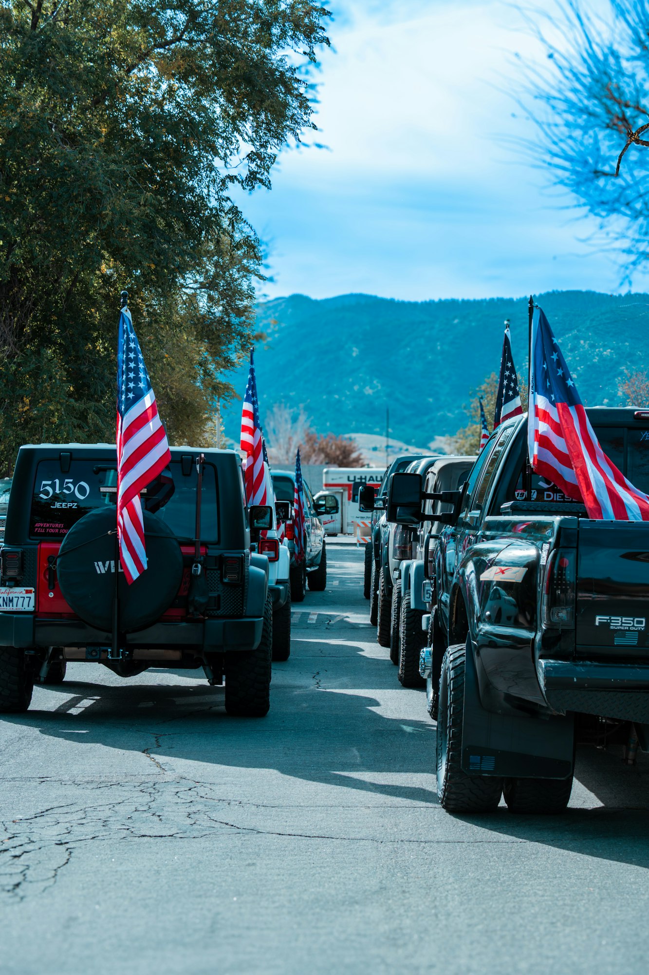 A line of vehicles with American flags, parked on a road surrounded by trees and mountains.