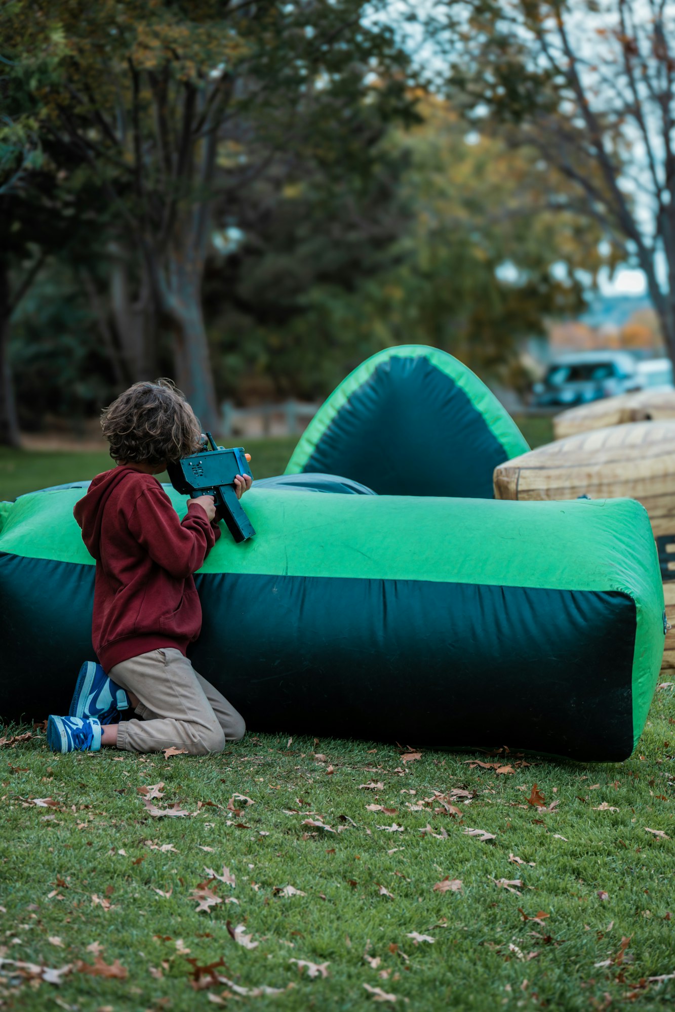 Child kneeling with toy gun, hiding behind an inflatable barrier in a park.