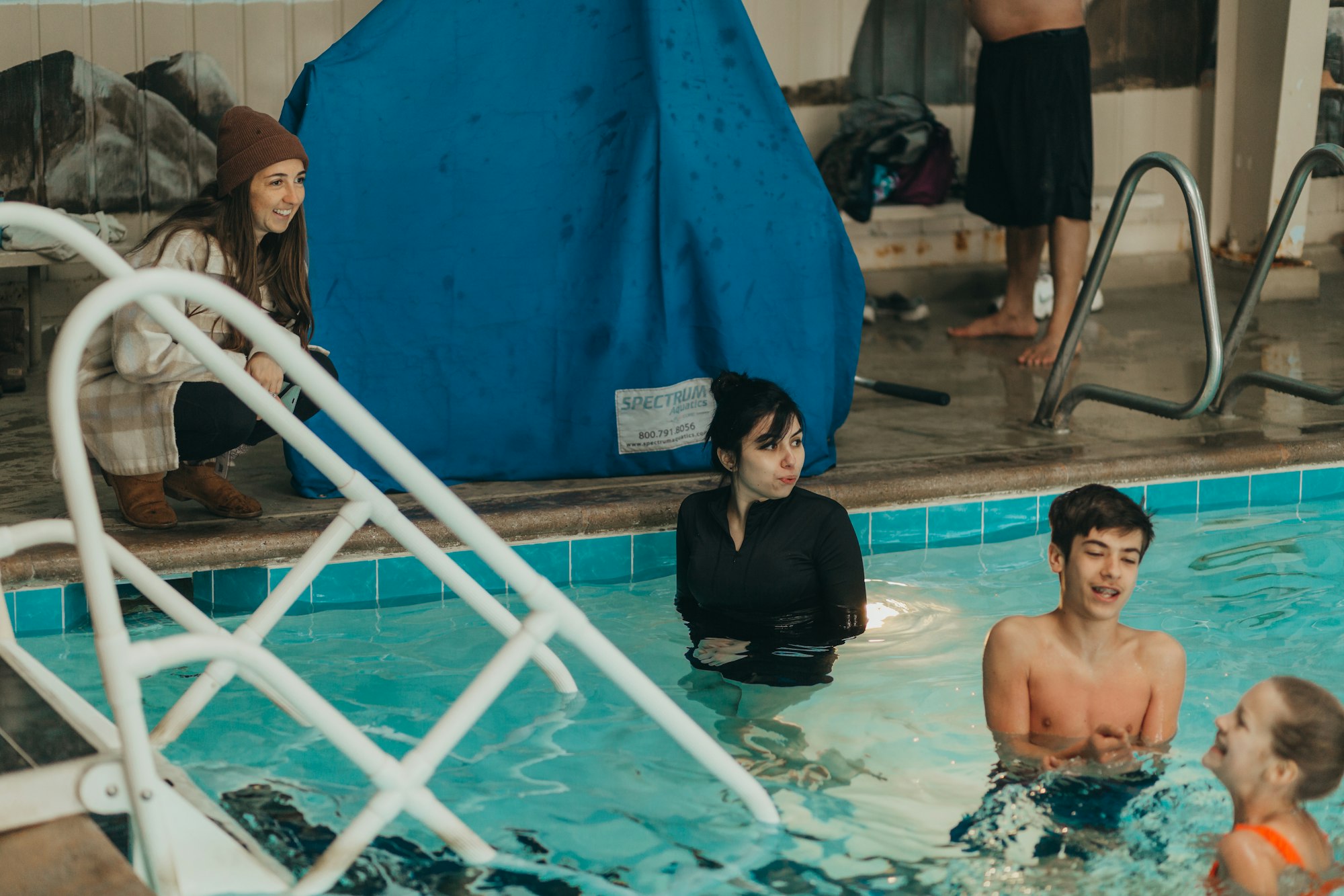 People enjoying time around an indoor pool, with a woman sitting by the edge and others in the water.