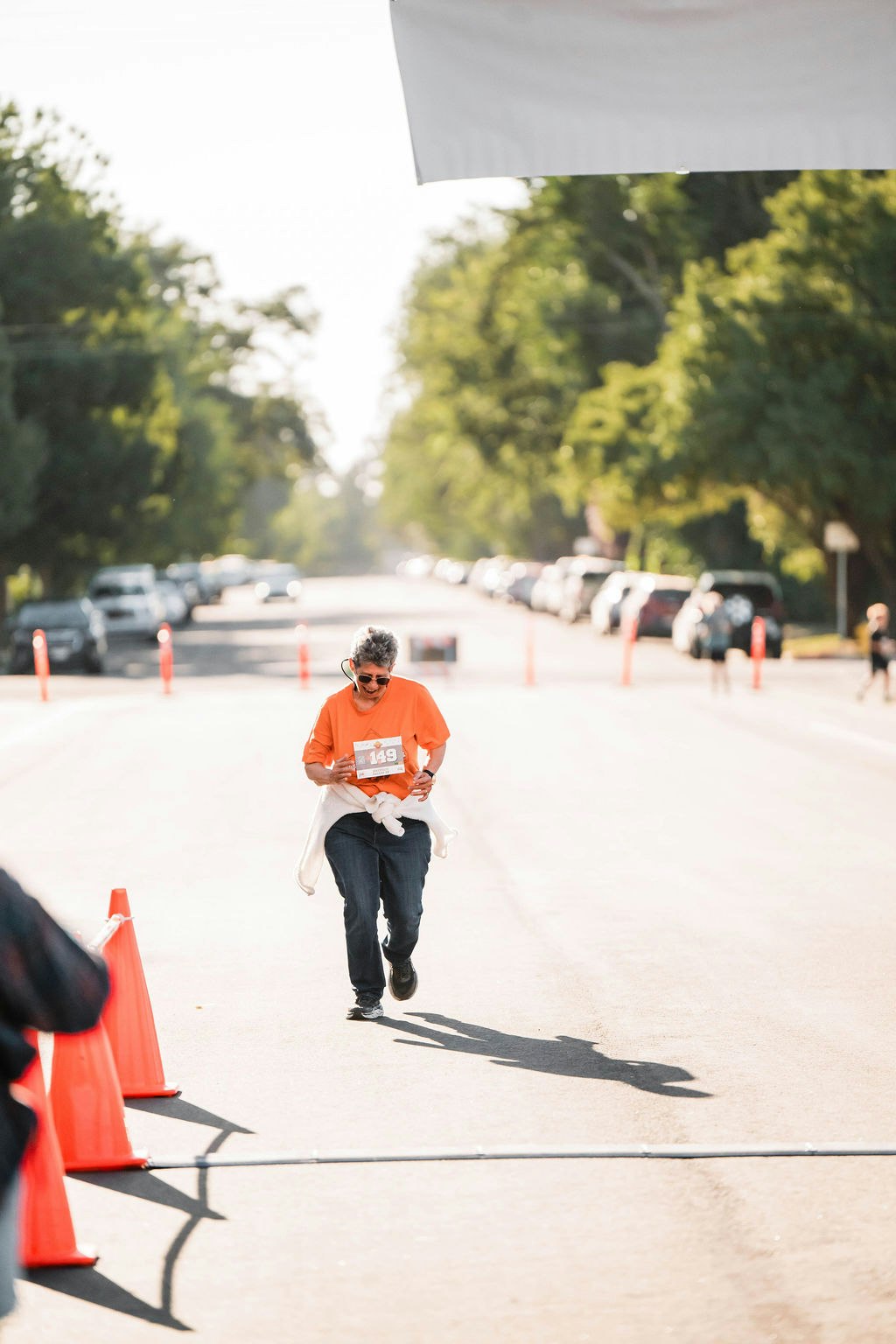 A person wearing an orange shirt and holding a race bib runs on a street with orange cones, trees, and parked cars in the background.