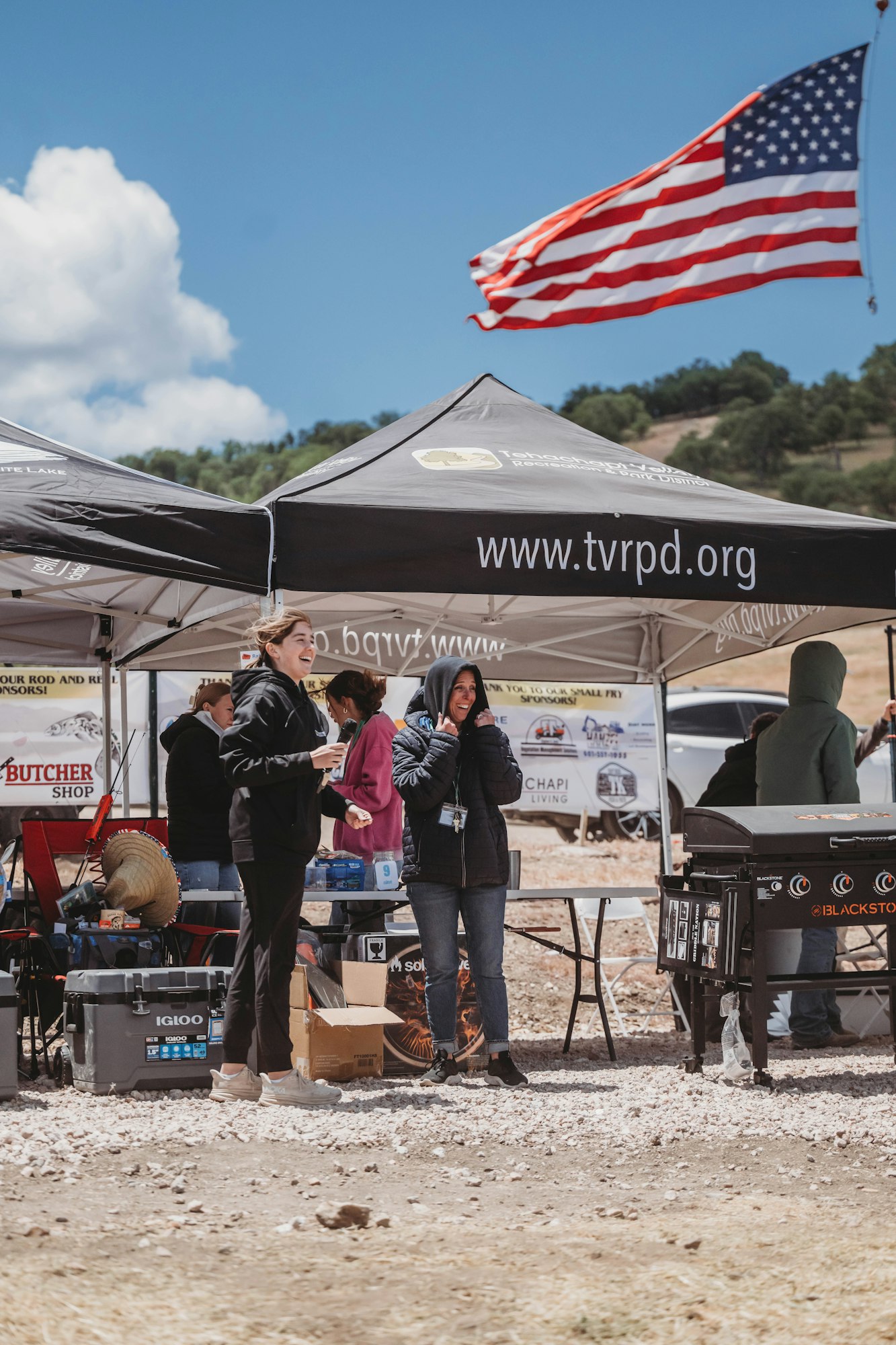 People at outdoor event by tents, with an American flag in the background; a website URL is visible on a tent.