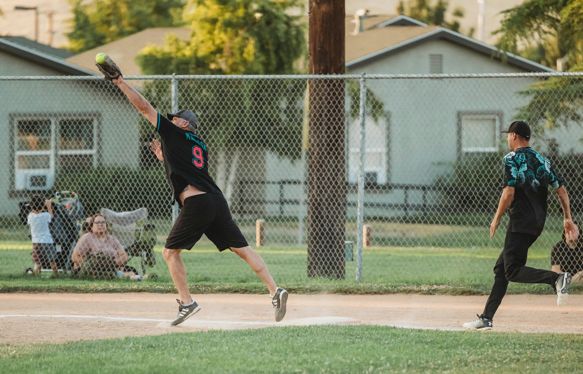 Men playing softball, one reaching for a catch, the other running bases, with spectators in the background.