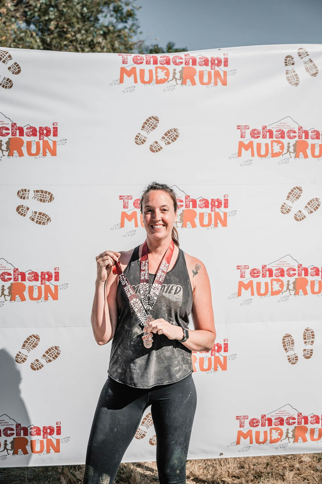 A woman holds medals in front of a "Tehachapi Mud Run" banner, smiling after completing an event.