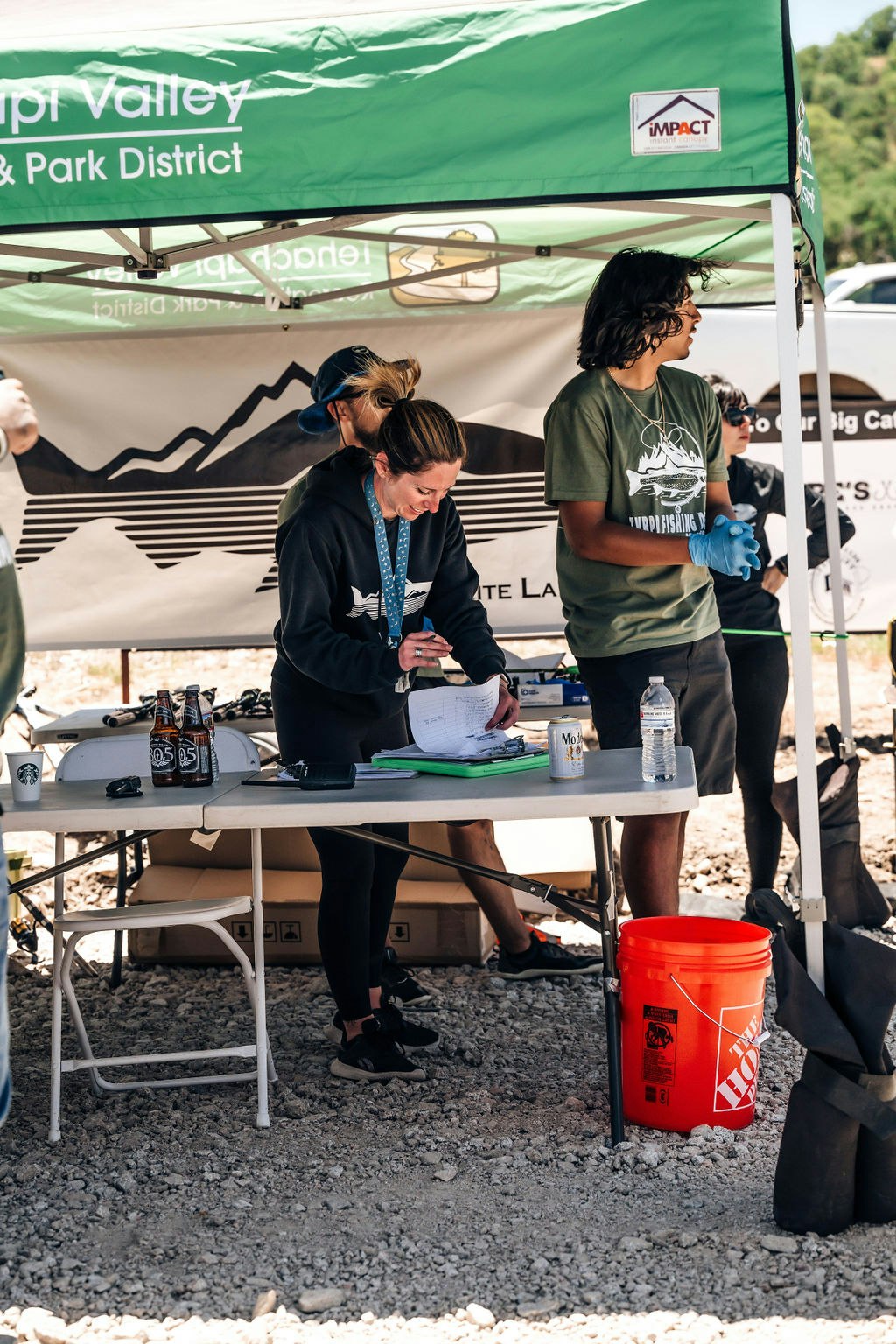 People under a tent with a banner, working on a table with papers and drinks, at an outdoor event.