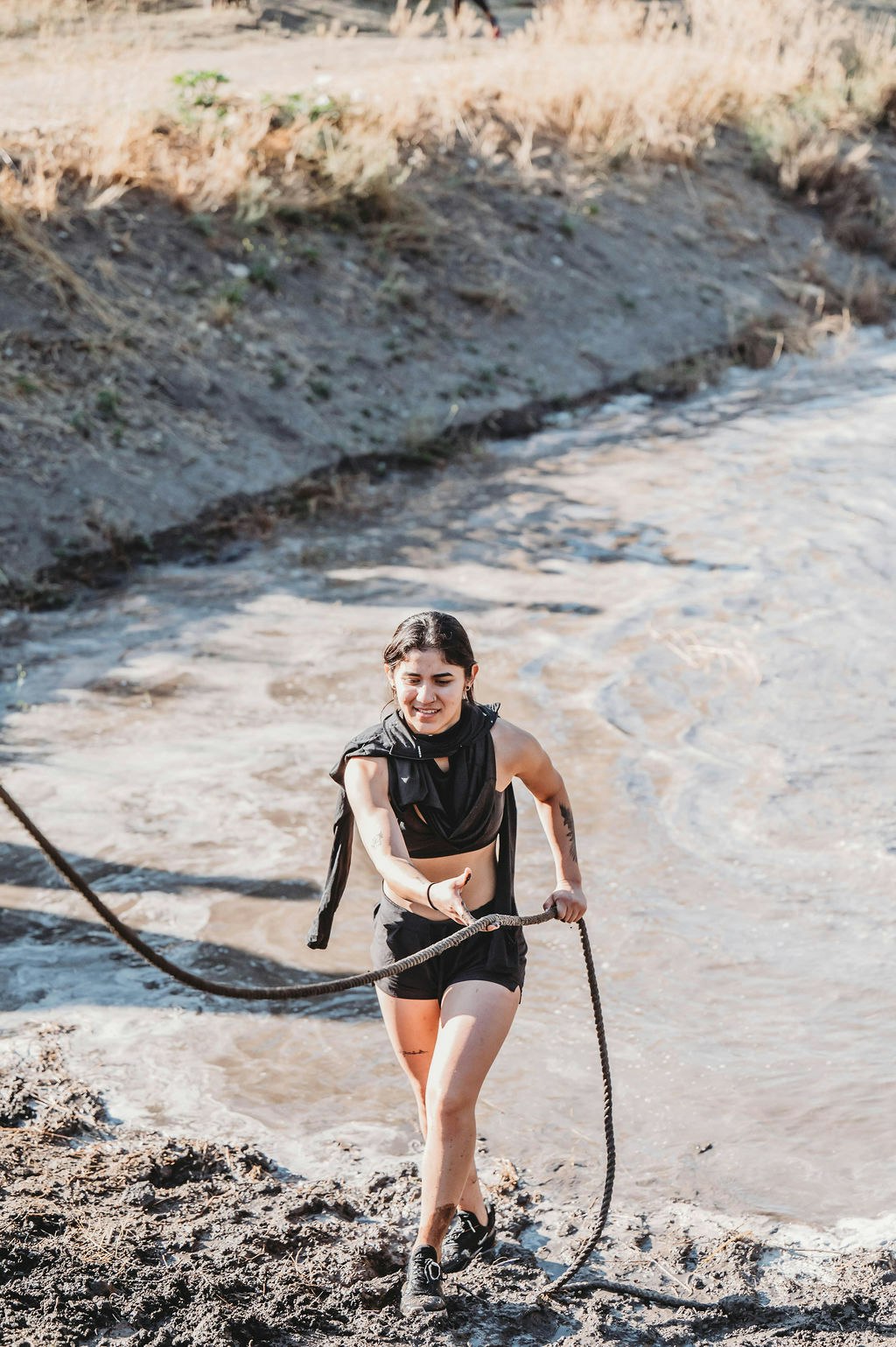 Person in athletic wear pulling a rope through a muddy obstacle course, smiling and focused.