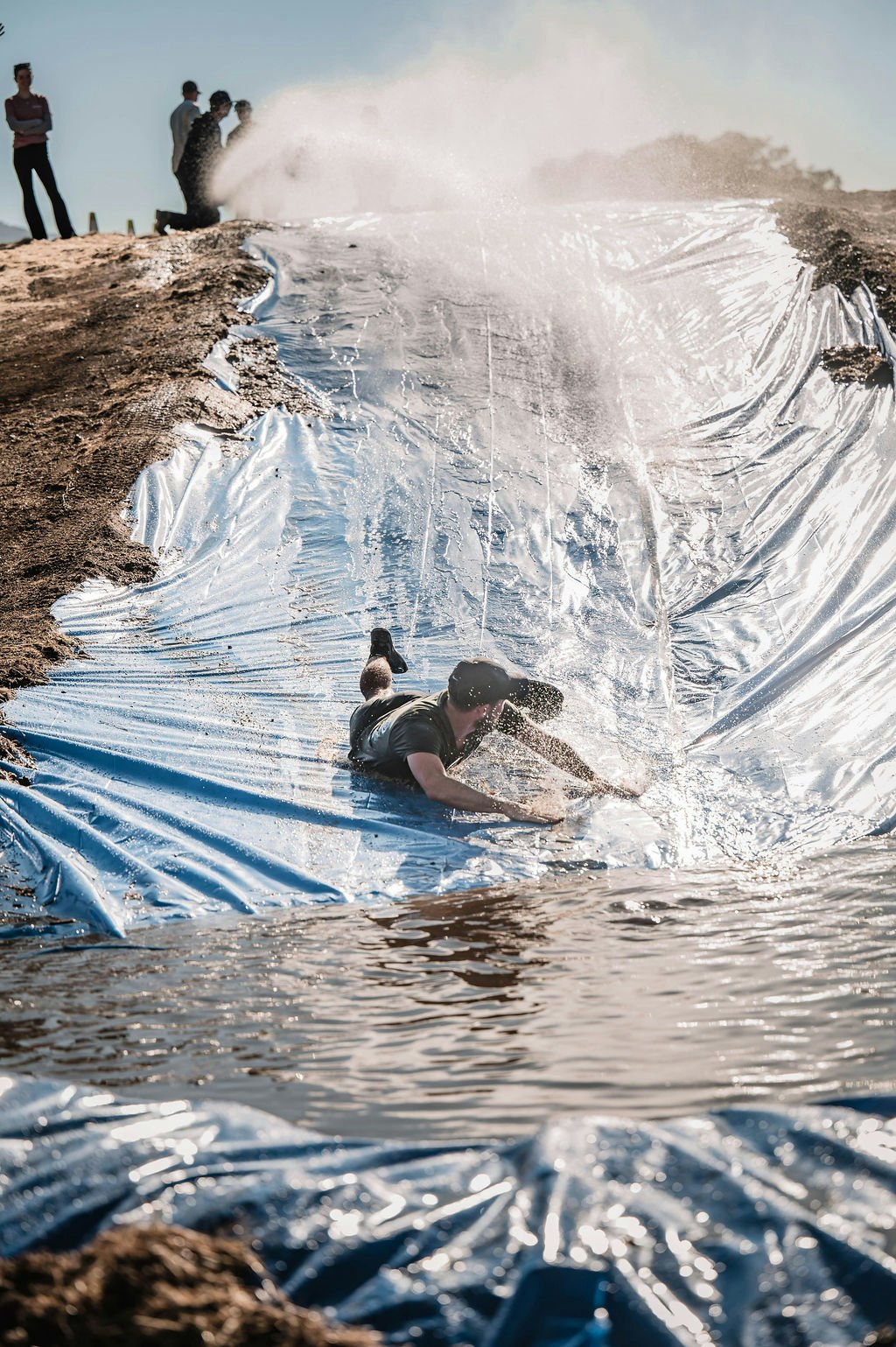 A person sliding down a water slide made of blue tarp, splashing into water at the bottom, while others watch.