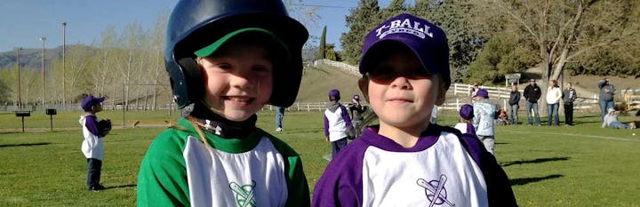 Two young children in baseball gear smile for the camera at a T-ball event, surrounded by other players and spectators.