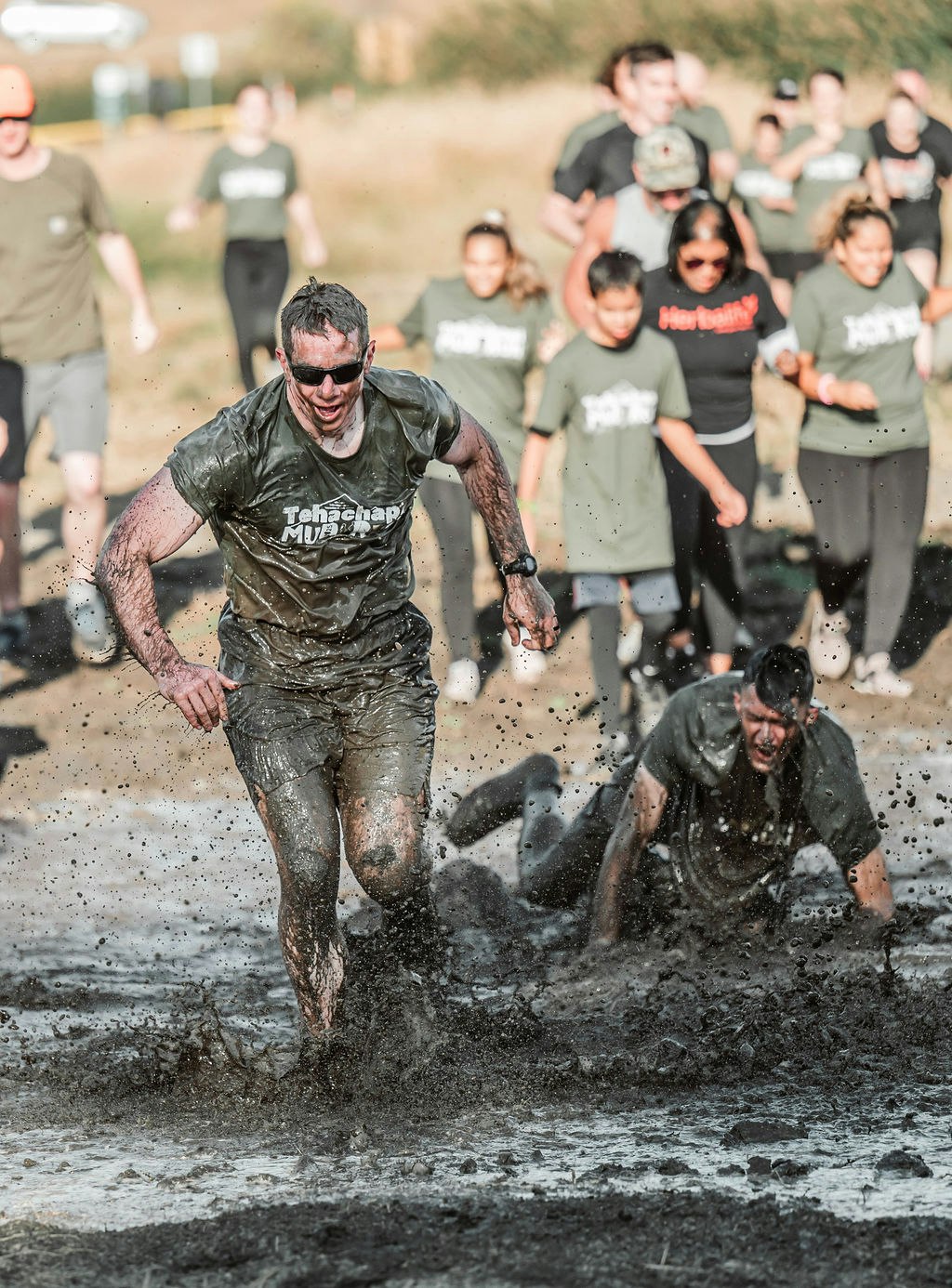 People participating in a muddy obstacle race, one running and another falling in the mud.