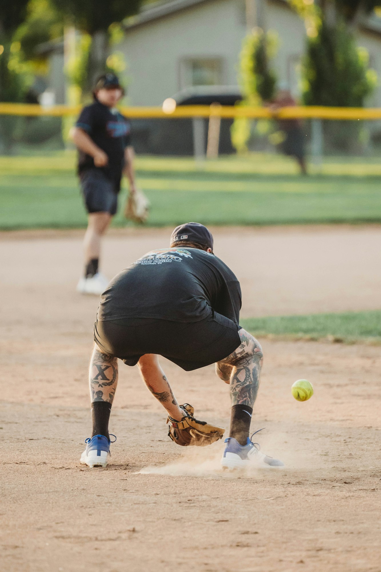 A softball player fielding a ball on a dusty infield, with another player in the background.