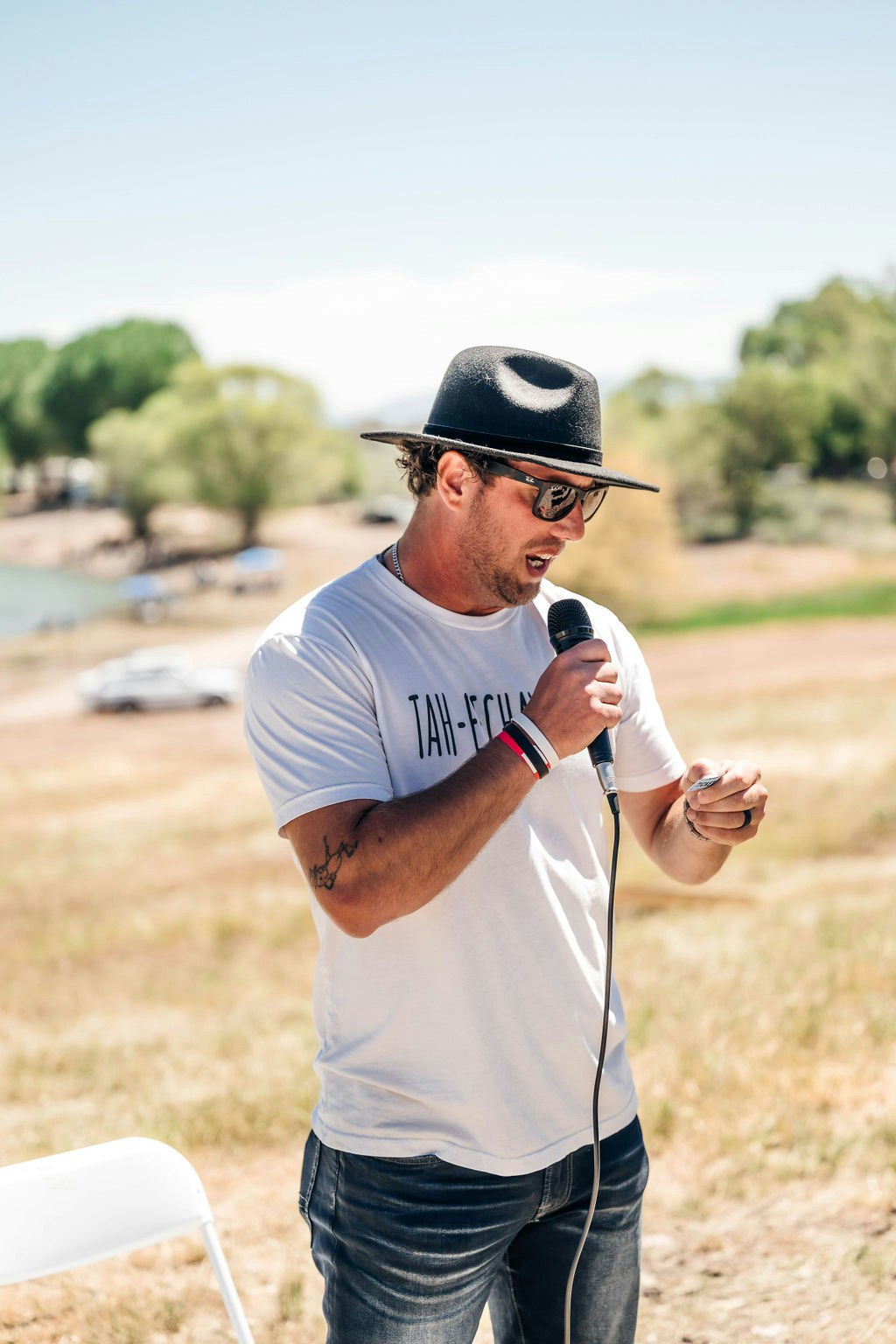 Man in hat and sunglasses speaking into a microphone outdoors.