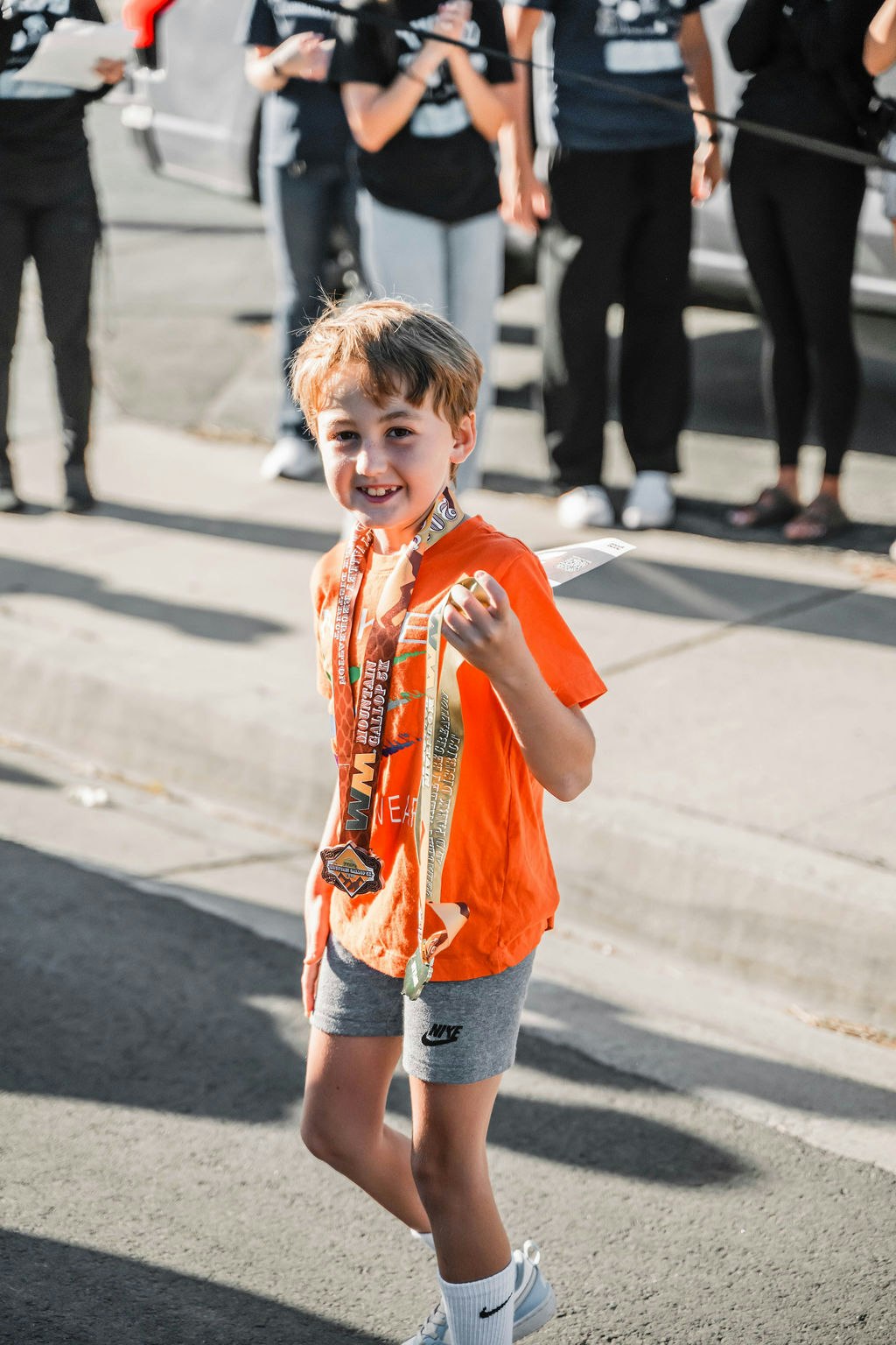 A smiling child in an orange shirt, holding medals around the neck, stands gleefully with people in the background.