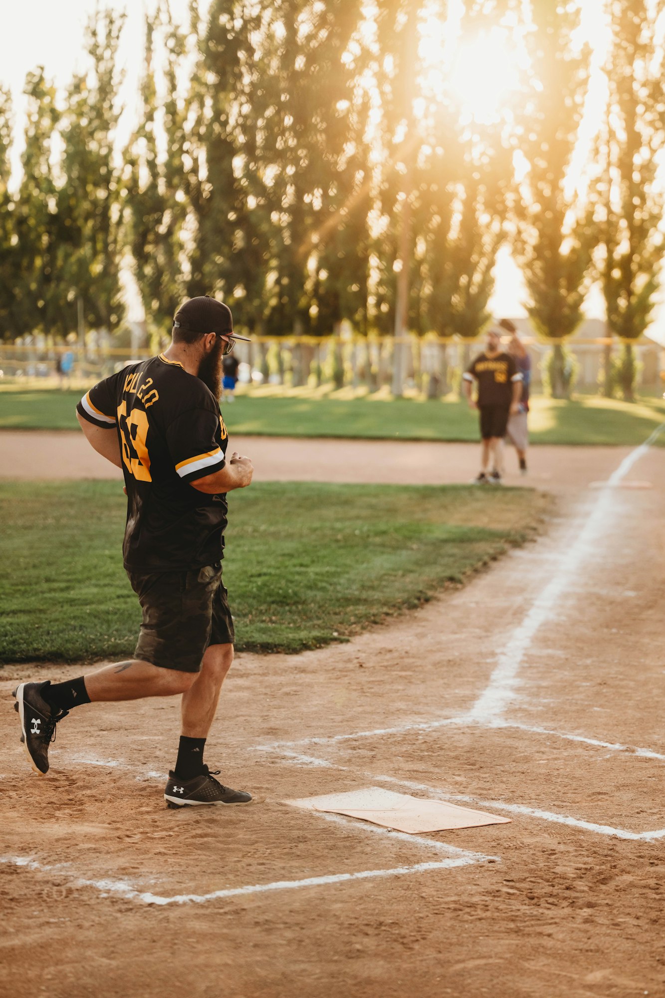 A player running bases on a baseball field at sunset, with other players and trees in the background.
