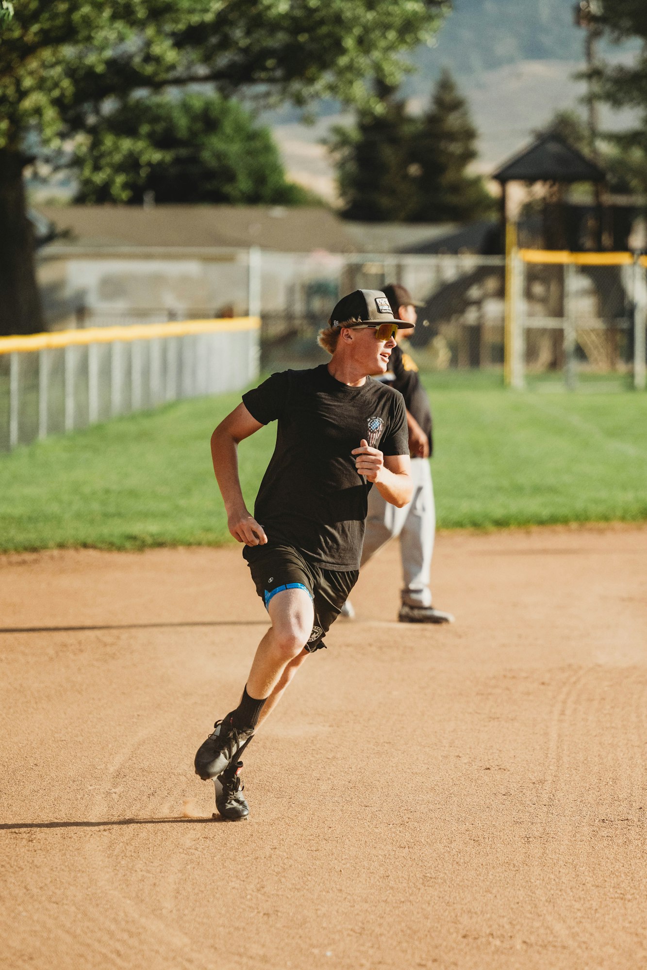 A person running on a baseball diamond, clad in casual sportswear and sunglasses, with trees and fences in the background.