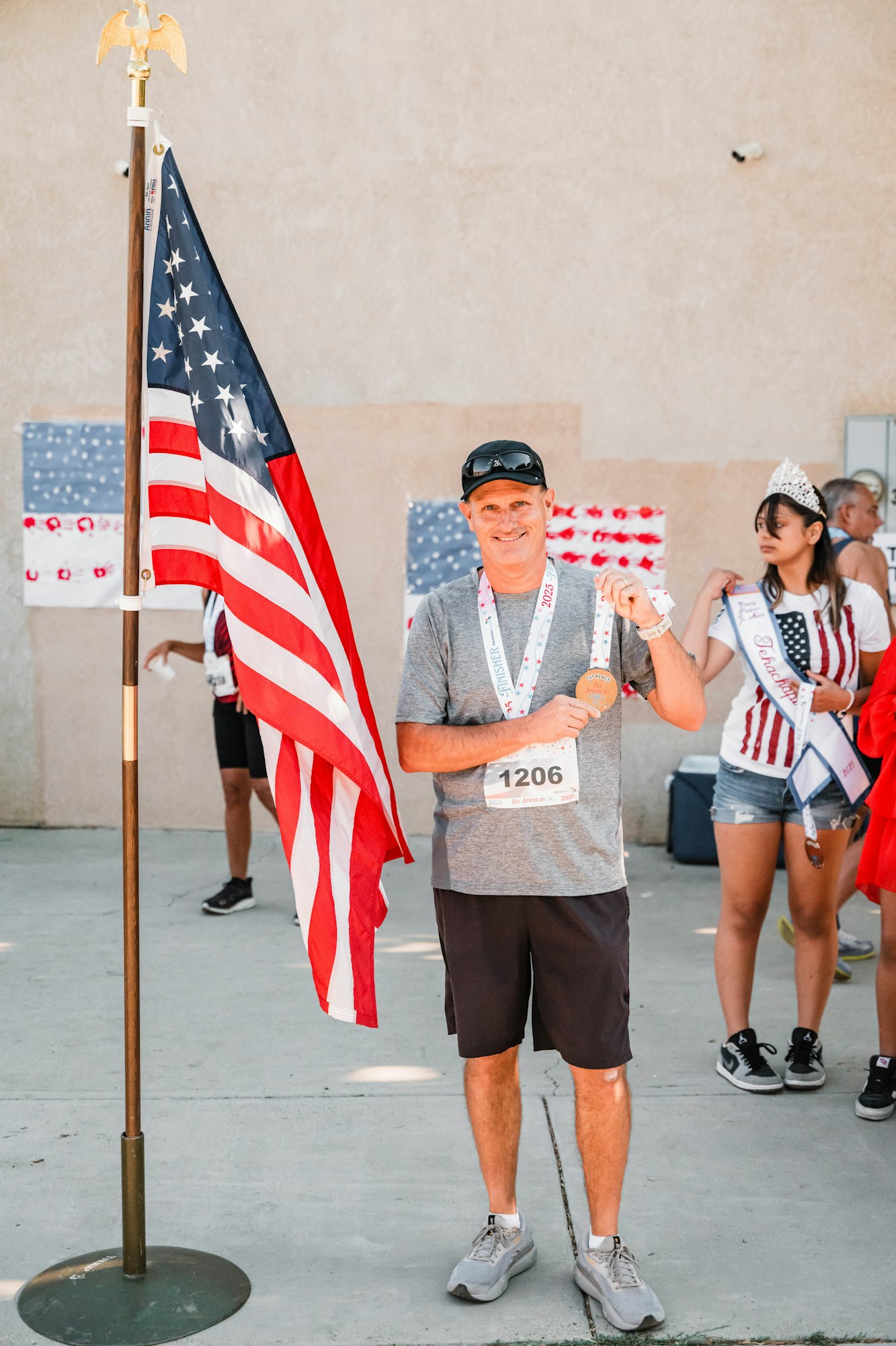 A person holding a medal stands beside a U.S. flag, with others in patriotic attire in the background.