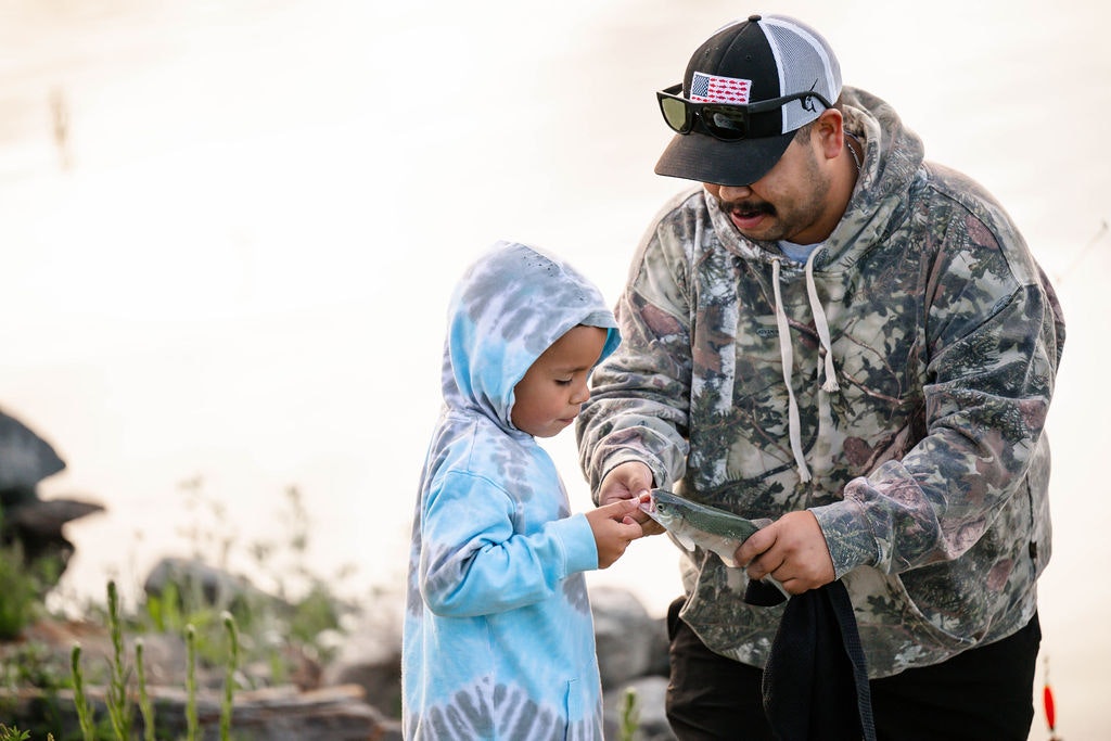 A man and a child in hoodies examine a fish outdoors.