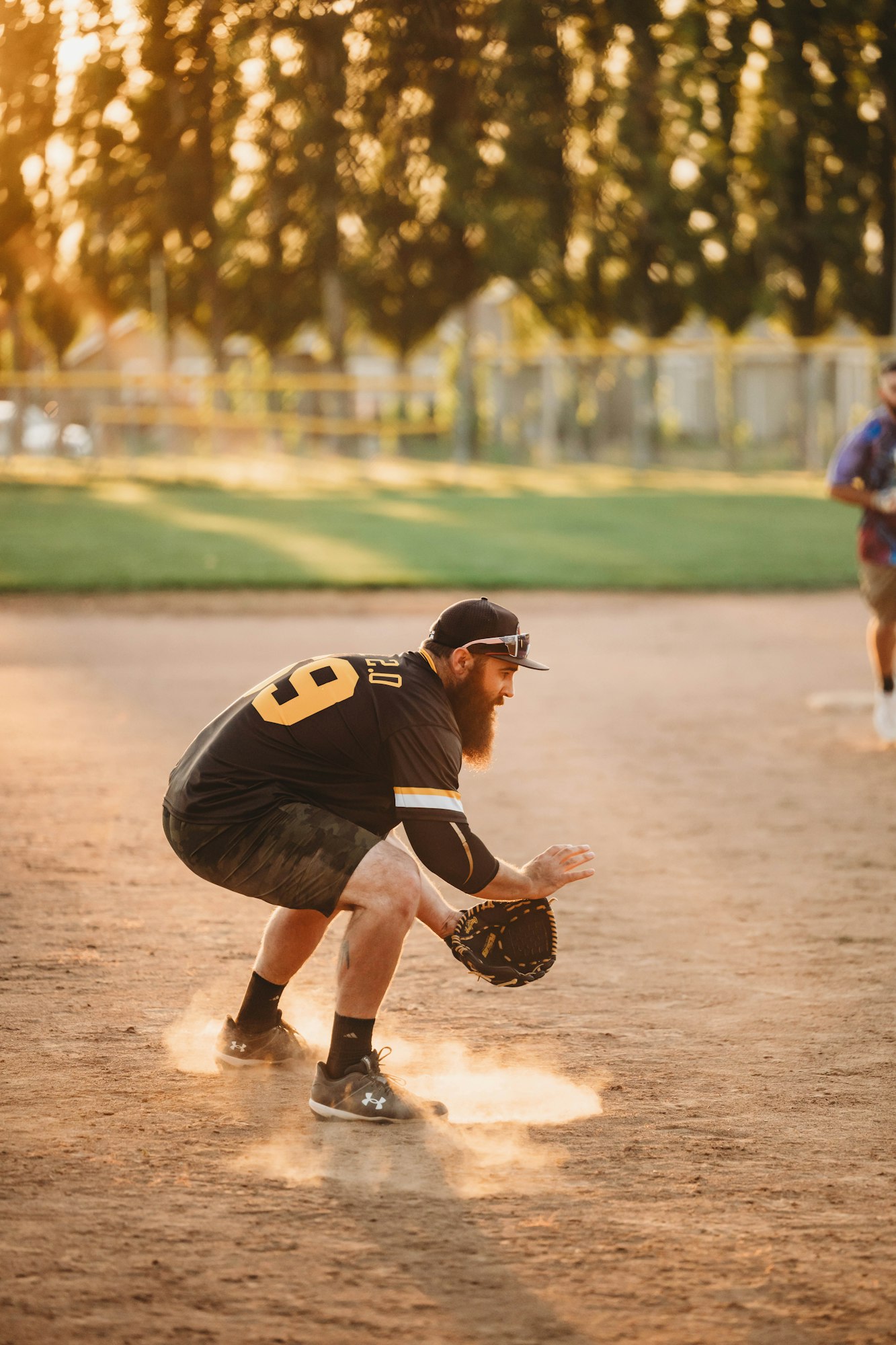 A baseball player crouching on the field with a glove, dust rising around his feet, trees and sunlight in the background.