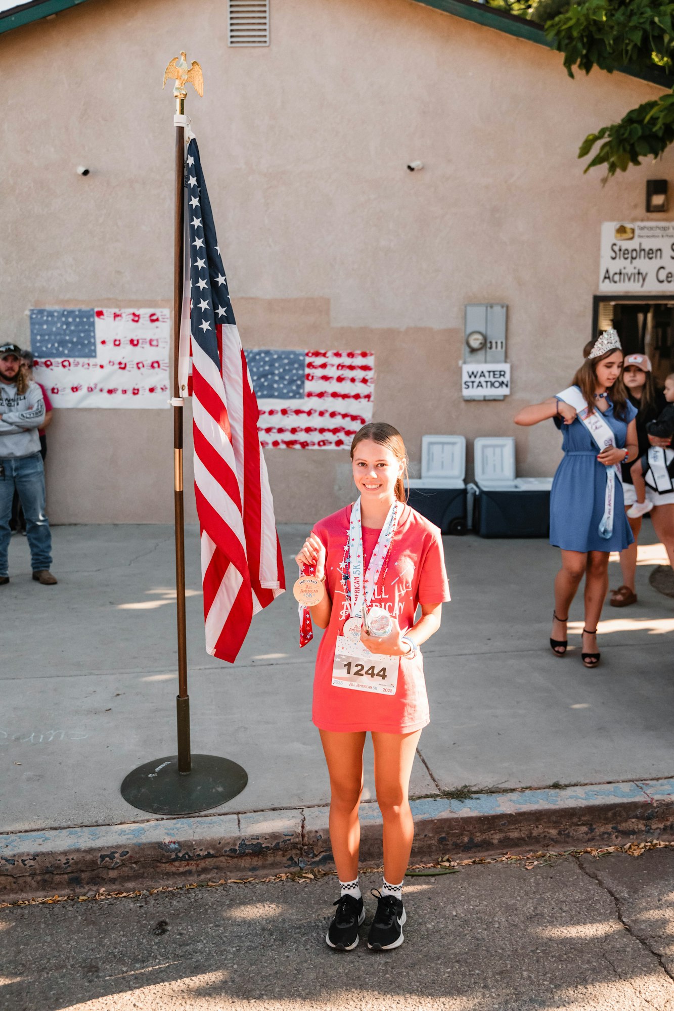 A girl with medals stands next to an American flag at an event.
