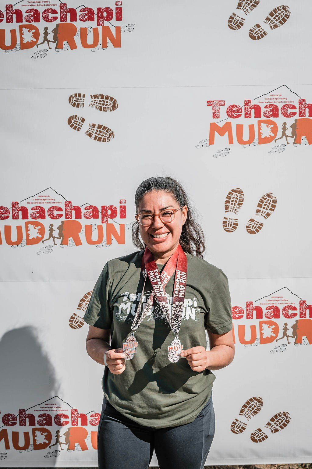 A person smiles, holding medals, in front of a "Tehachapi Mud Run" backdrop.
