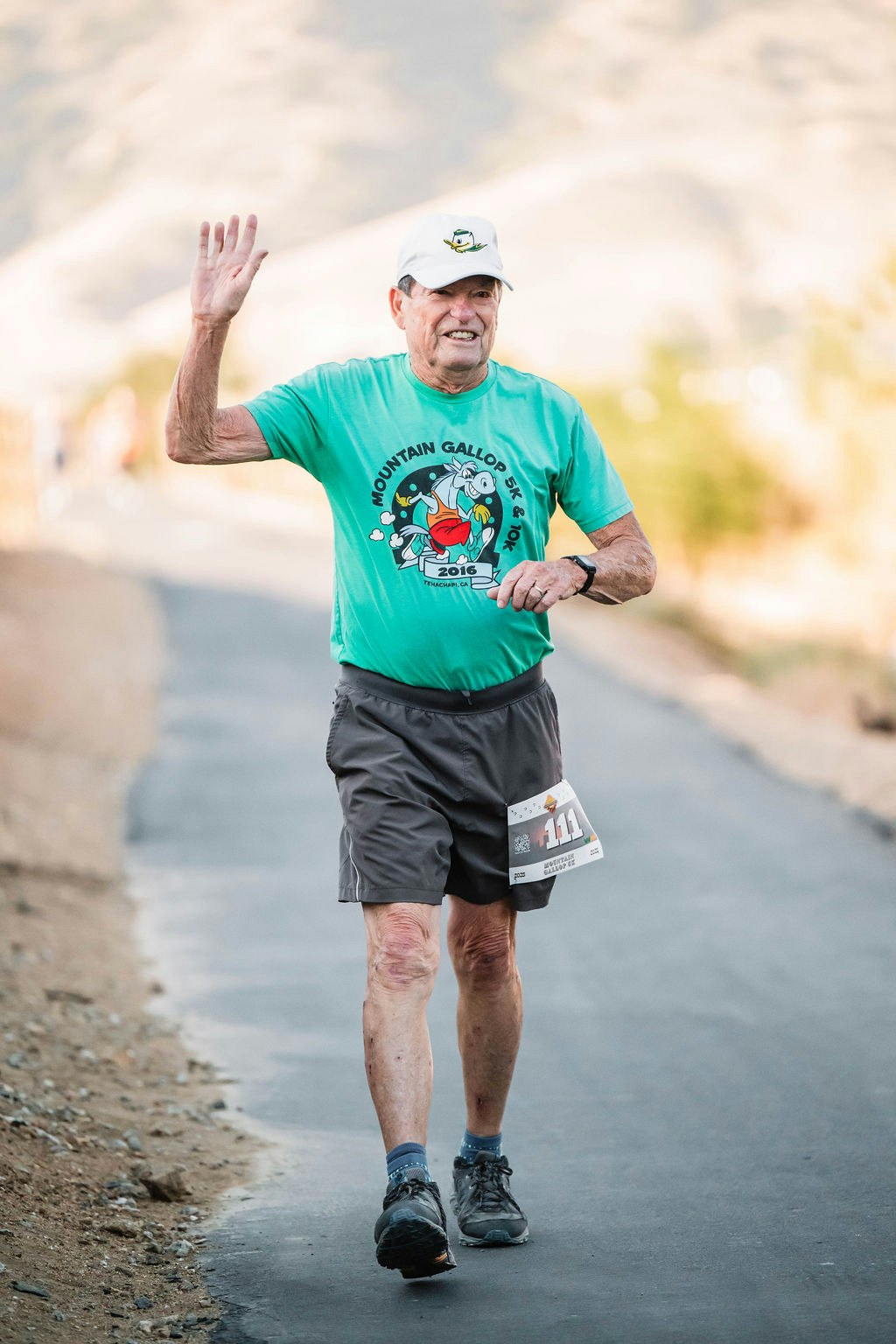 A person running on a path, wearing a green "Mountain Gallop 5K & 10K" T-shirt and a hat, with a race bib number 111.