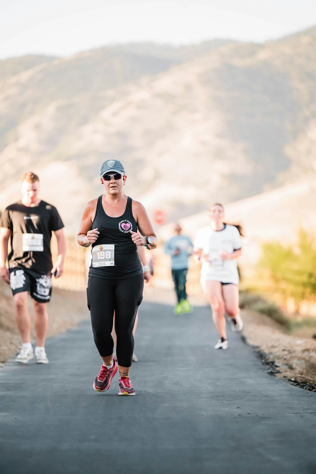 People running outdoors on a paved path, wearing athletic gear and race numbers, with a hilly landscape in the background.