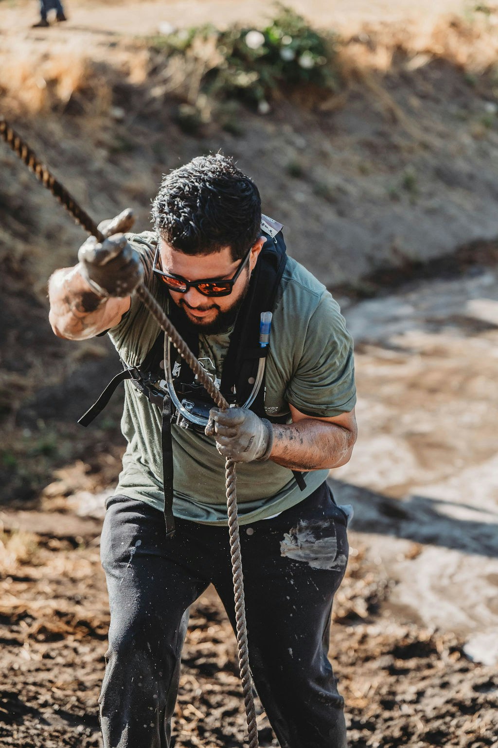 Person climbing a muddy hill using a rope, wearing sunglasses and gloves.