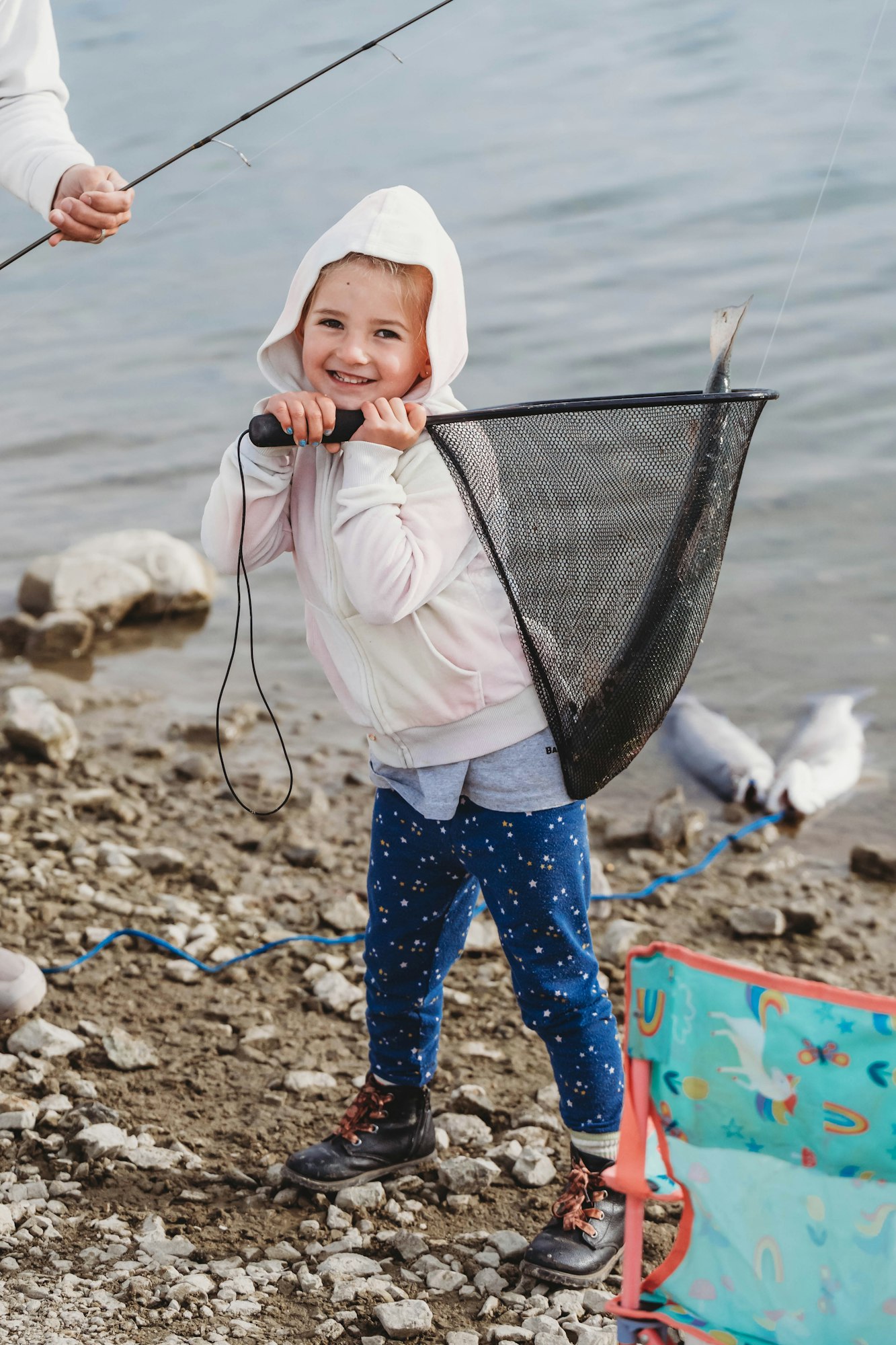 A child in a hoodie happily holds a fishing net with a fish, standing on a rocky shore.