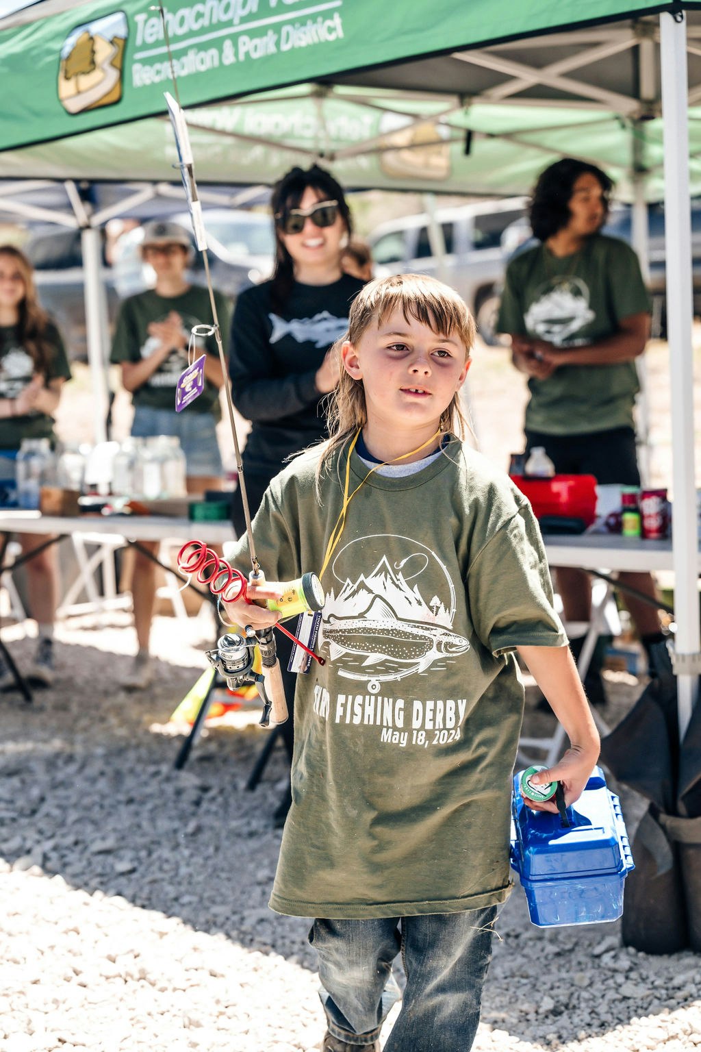 A child with a fishing rod, trophy, and tackle box at a fishing derby event.