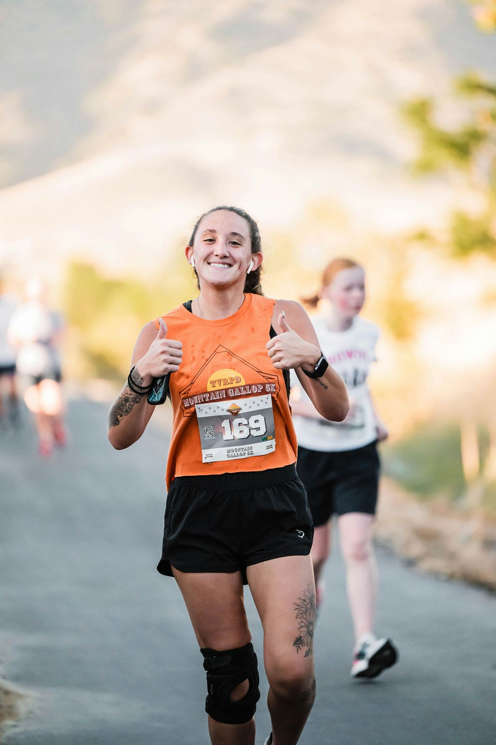 A smiling runner gives thumbs up during a race, wearing an orange shirt and black shorts, with a knee brace and race bib number 169.
