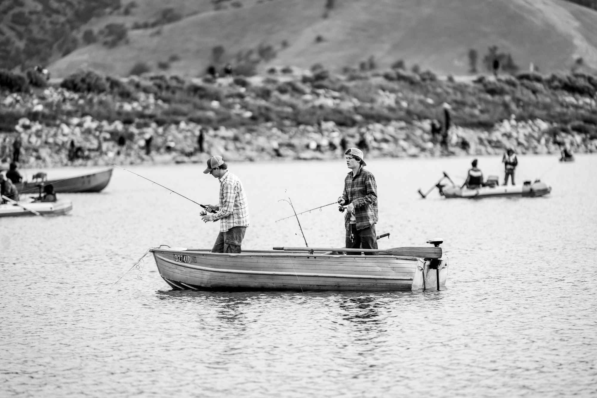 Two people fishing on a small boat in a lake, with hills and other boats in the background.