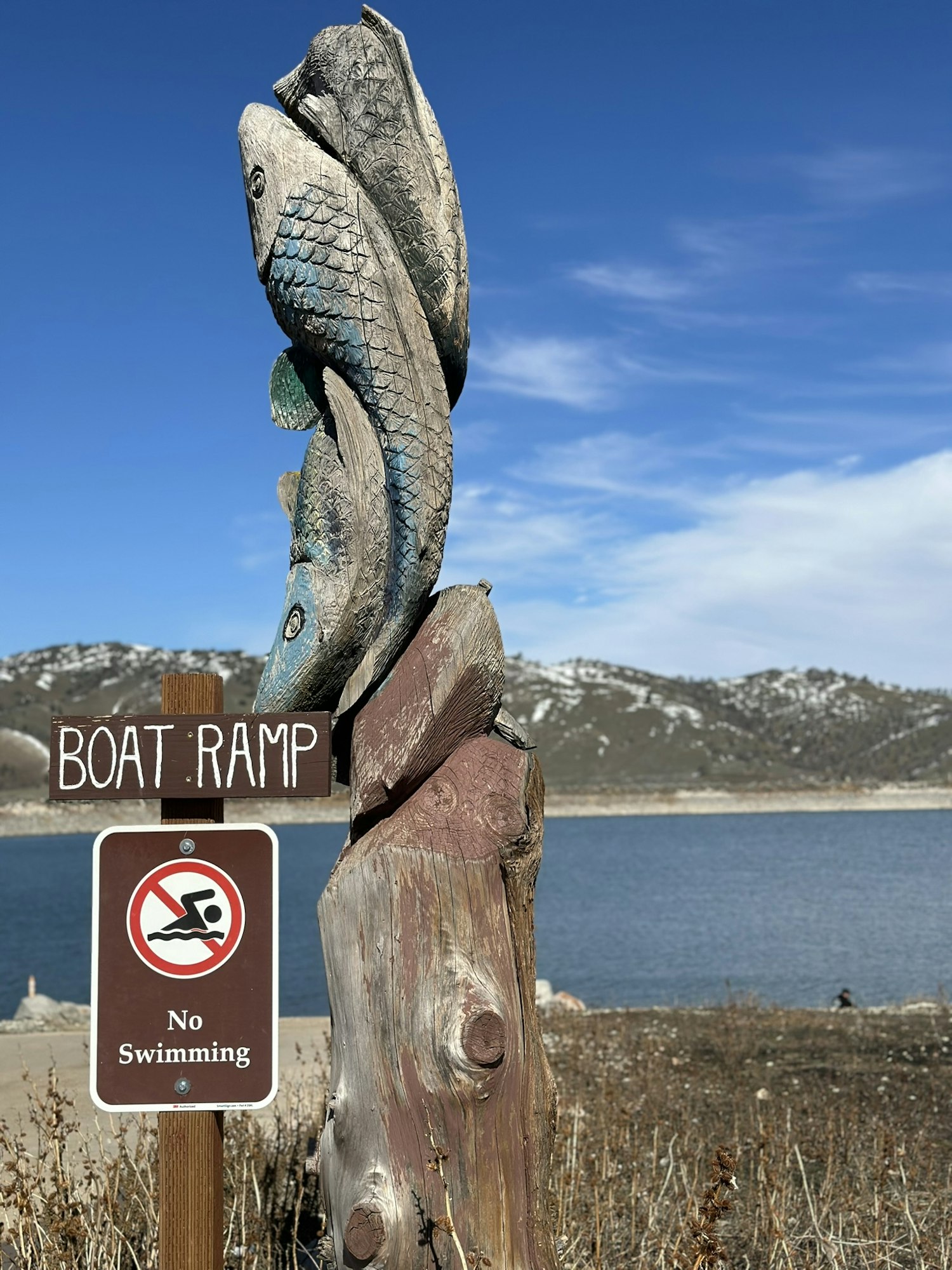 A wooden fish sculpture by a lake with a "No Swimming" sign and "BOAT RAMP" written on a post.