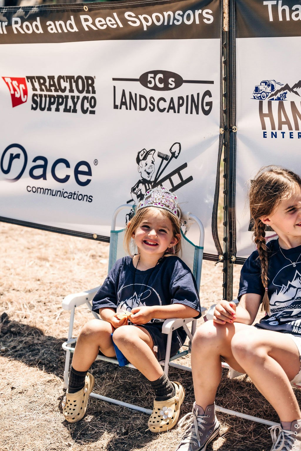 Two smiling kids sitting outdoors, one wearing a tiara, with a banner of logos behind them.