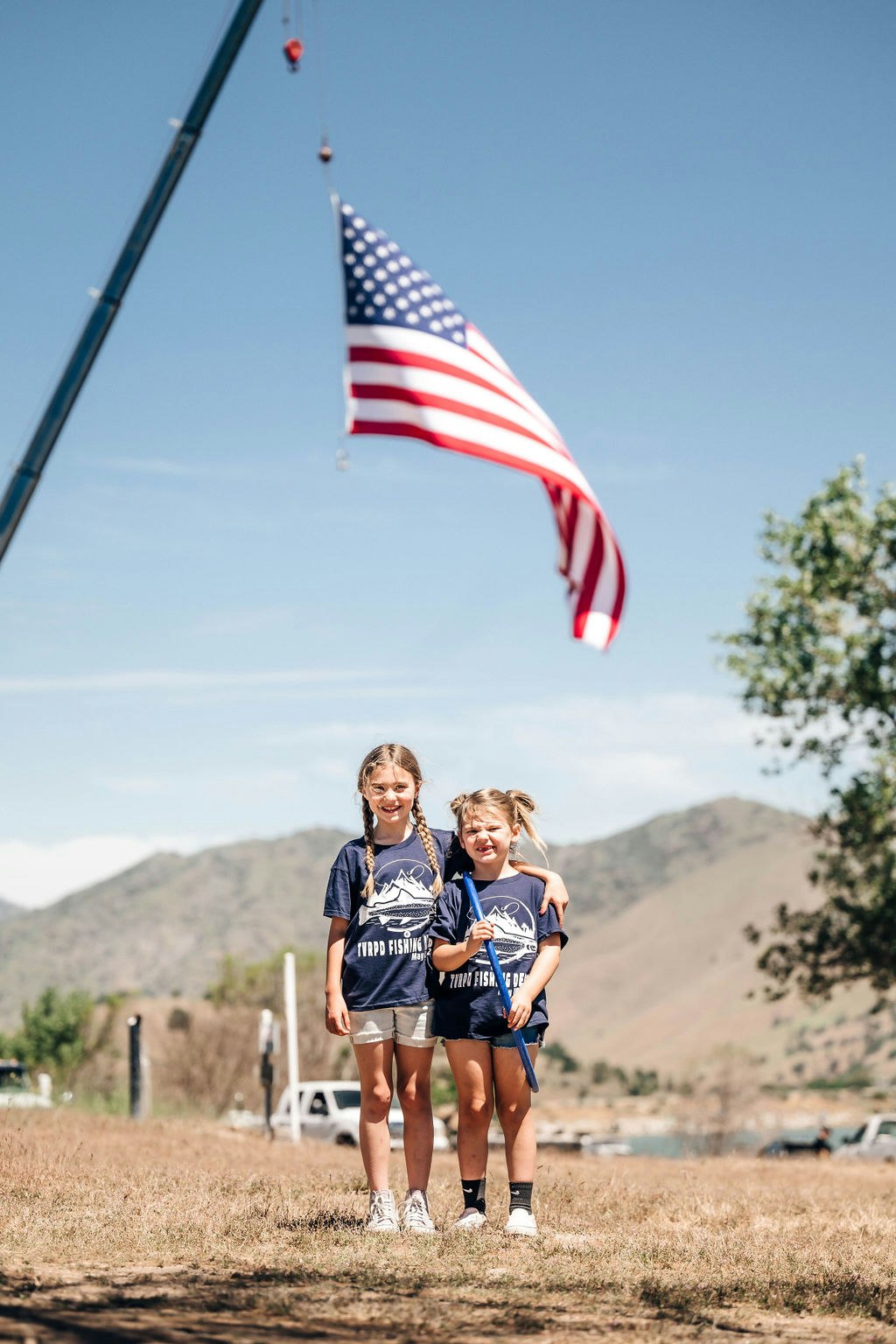 Two children smiling outdoors under a large American flag hanging from a crane, hills in the background.