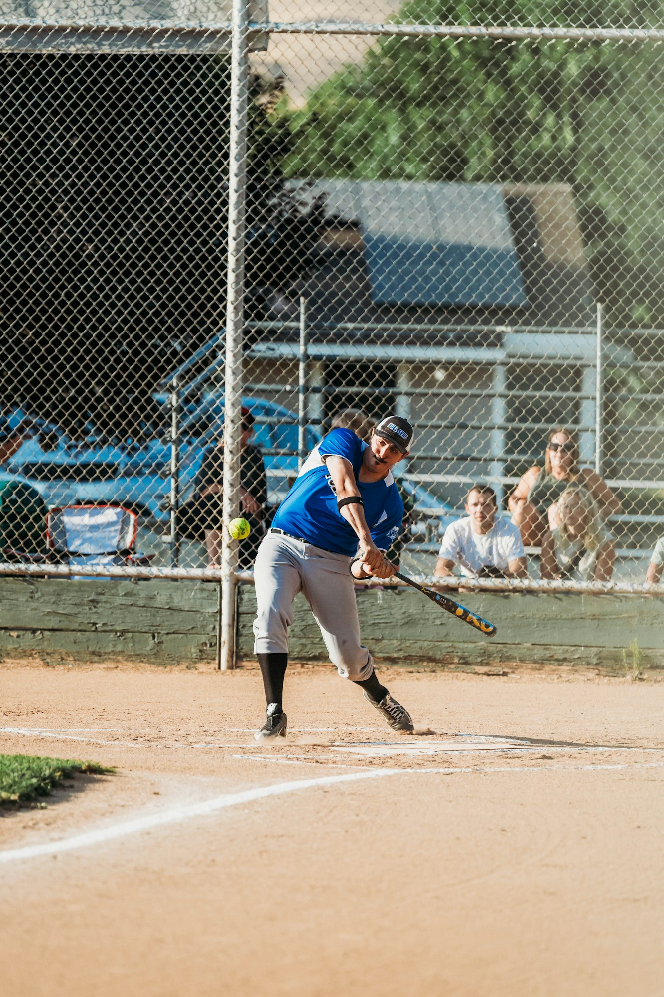 A baseball player swinging a bat at a ball on a sandy field with a fence and spectators in the background.