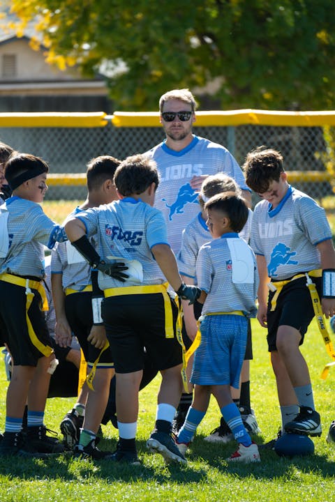 A youth flag football team huddles on a field with their coach, wearing matching uniforms.