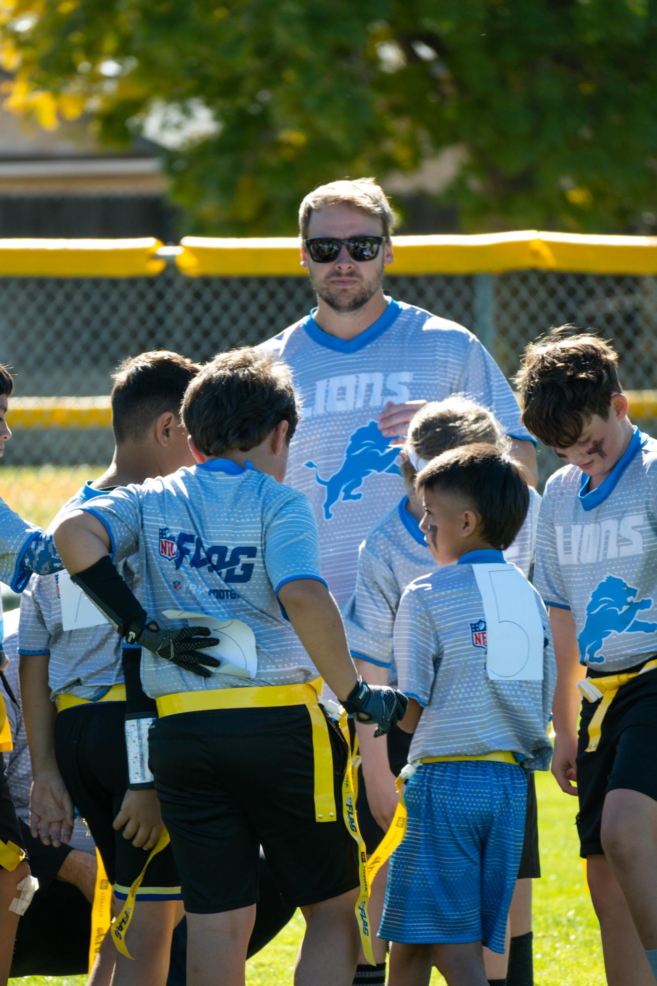 A youth flag football team huddles on a field with their coach, wearing matching uniforms.