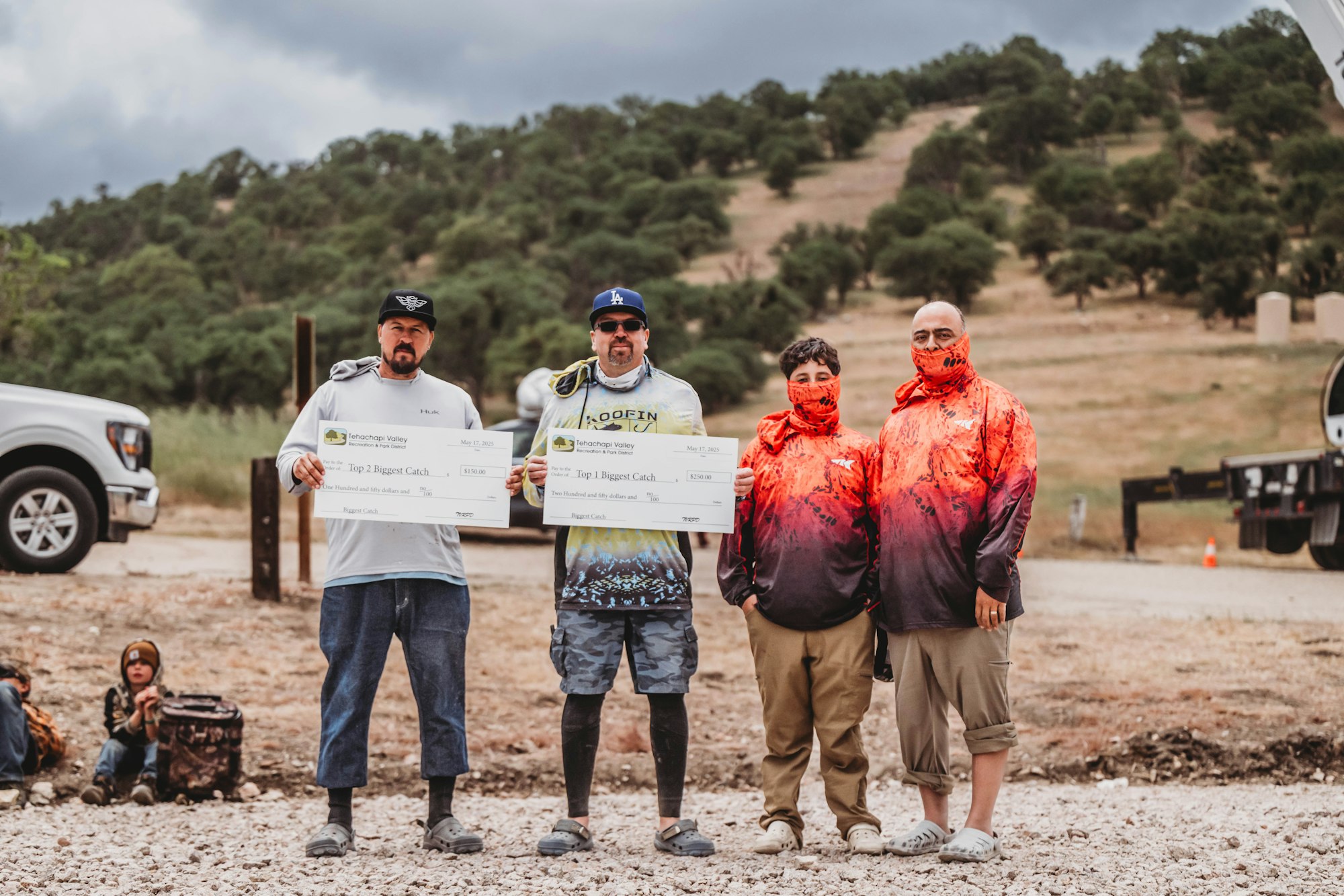 Four people outdoors, two holding large checks labeled "Tehachapi Valley," wearing casual clothing and masks. Trees and hills in the background.
