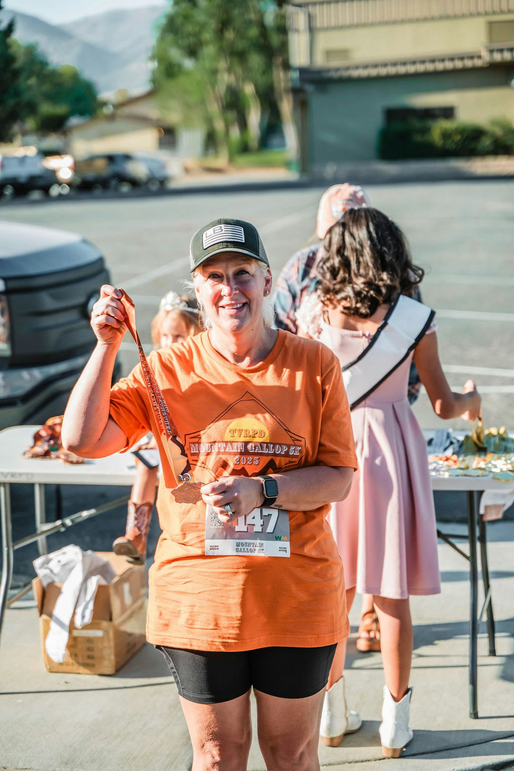 A person holding a medal, wearing an orange event shirt and a race bib, with people and tables in the background.