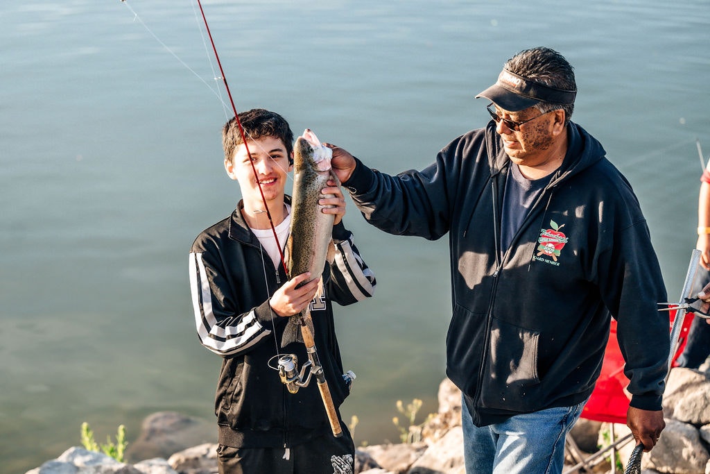 A young man holding a fish next to an older man by a lakeside, likely celebrating a successful catch while fishing.