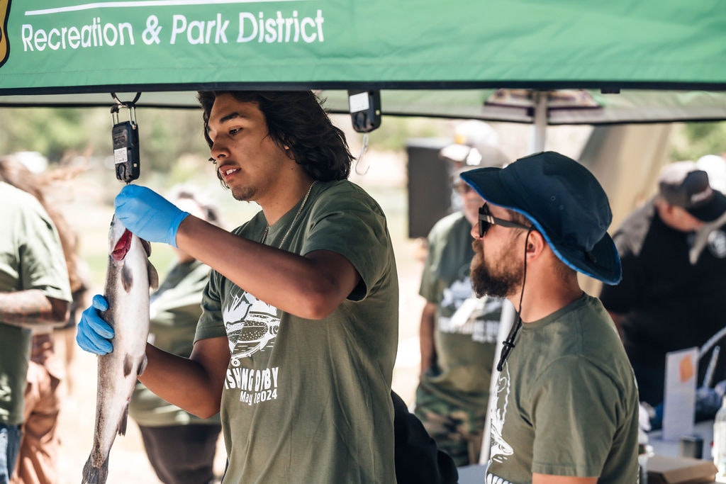 A person weighing a fish at an event, another person looking on, both under a "Recreation & Park District" tent.