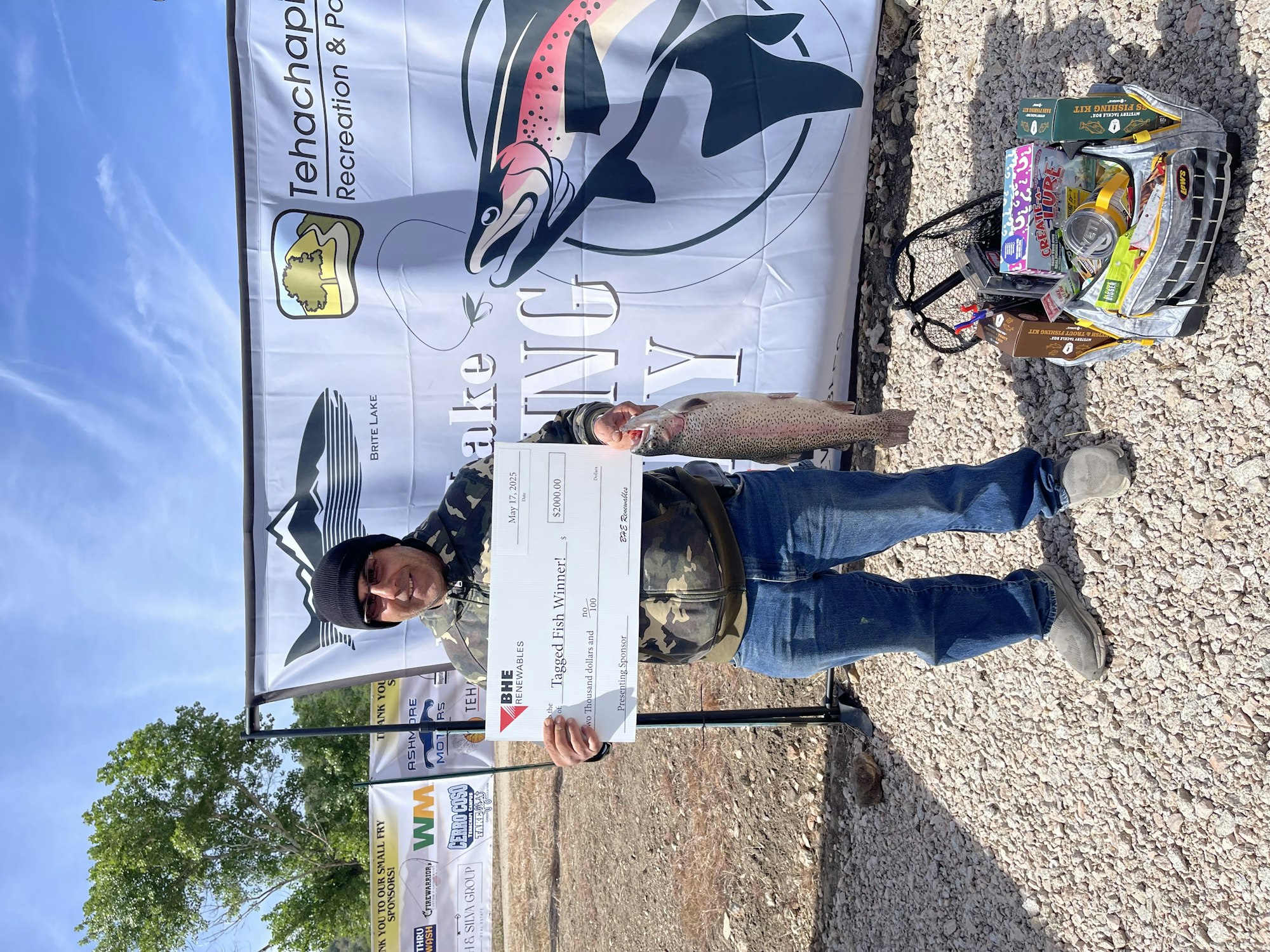 A person holding a large fish and a $500 check, with a fishing tournament banner and fishing gear in the background.