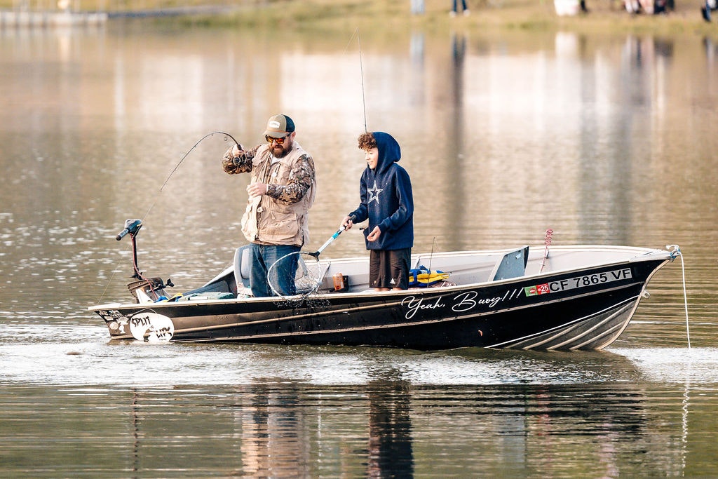 Two people on a boat, one fishing with a bent rod, the other watching, surrounded by calm water.