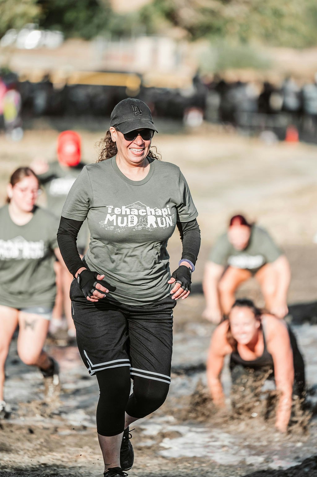 Participants in a mud run, wearing "Tehachapi Mud Run" shirts, are running and crawling through muddy terrain.