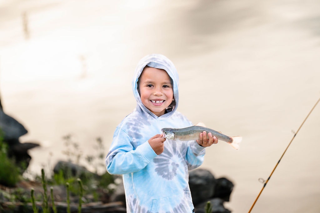 A smiling child in a hoodie holding a fish by a river, with a fishing rod nearby.