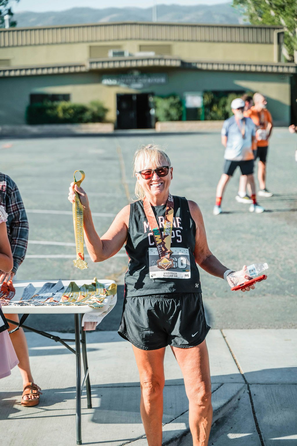 A person in sunglasses holds a medal and water, wearing a race bib, standing beside a table with more medals outside.