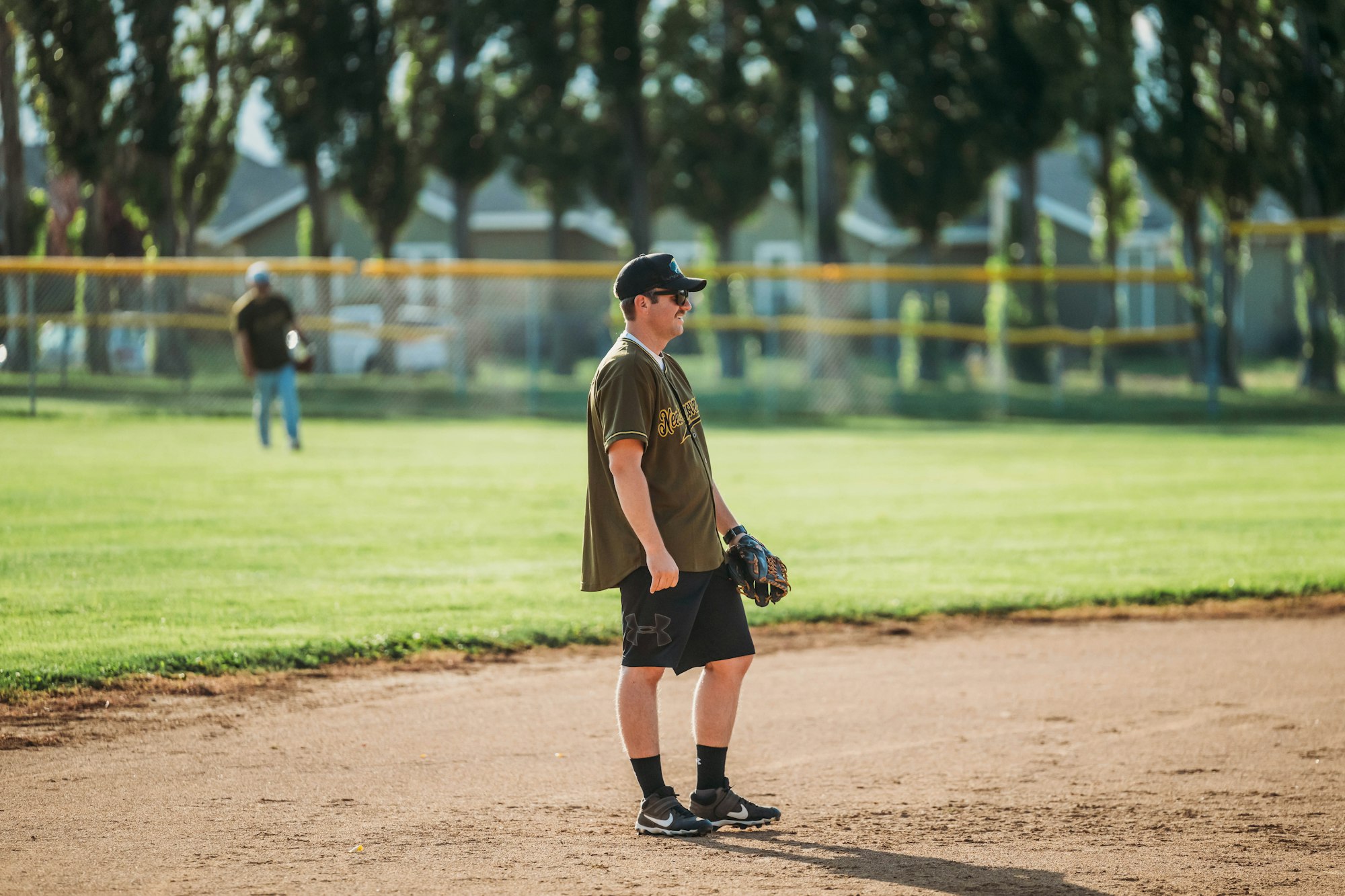 A person standing on a baseball field wearing a cap, t-shirt, shorts, and a glove. Trees and fence in the background.