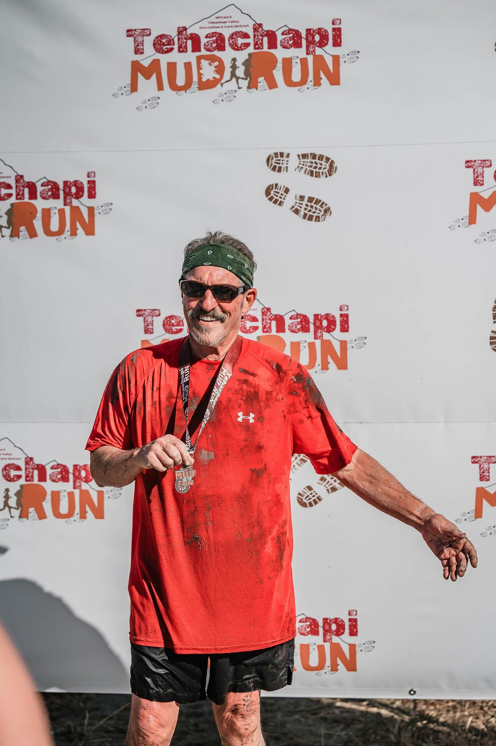 A man with a medal, wearing a red shirt, stands in front of a "Tehachapi Mud Run" banner, covered in mud and smiling.