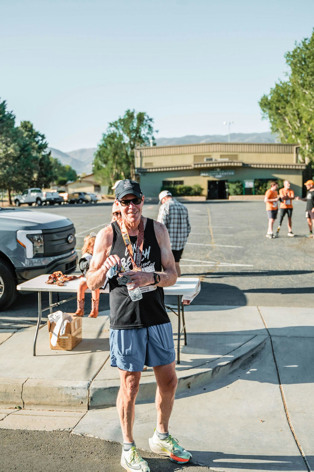 A person in athletic wear holds a medal and water bottle, standing near a table with people and a building in the background.