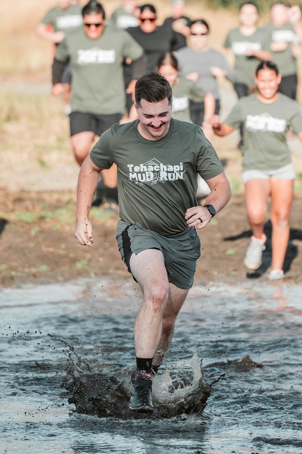 A man running through mud while participating in a "Tehachapi Mud Run" event, followed by other runners.