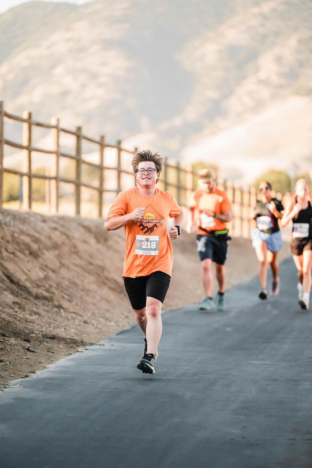 People running on a paved path in outdoor setting, wearing sports attire and numbered bibs, with mountains in the background.