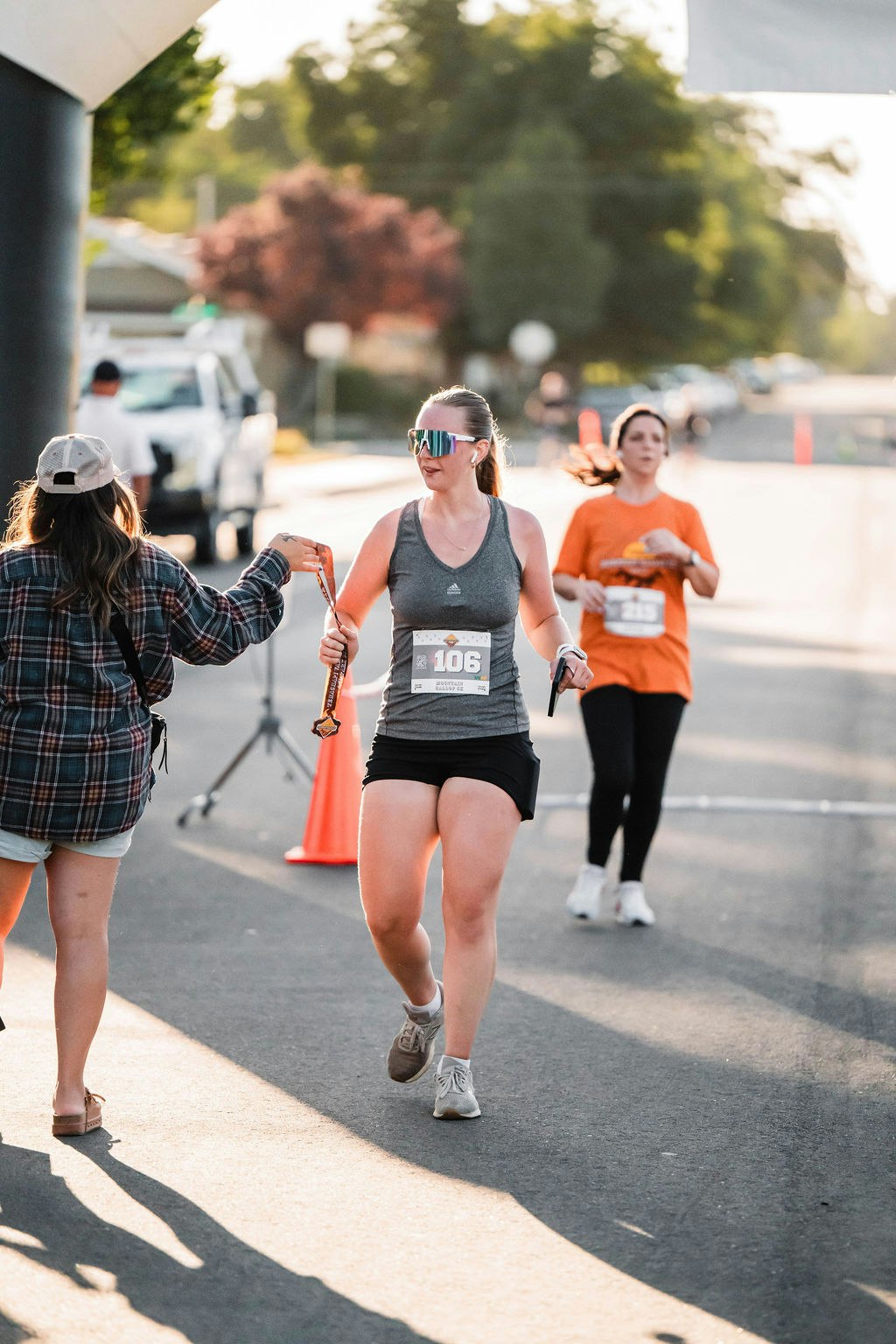 A runner with bib number 106 receives a medal from an onlooker during a race. Another runner follows in an orange shirt.